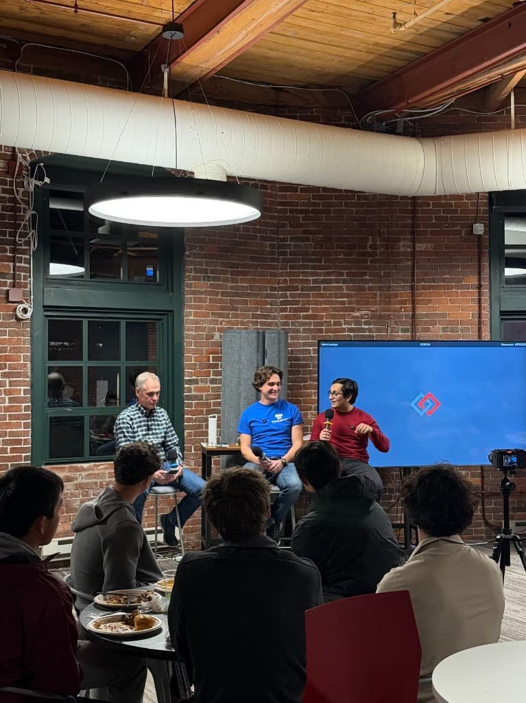 A panel discussion in a brick-walled loft event space with speakers on stools and an audience in the foreground.