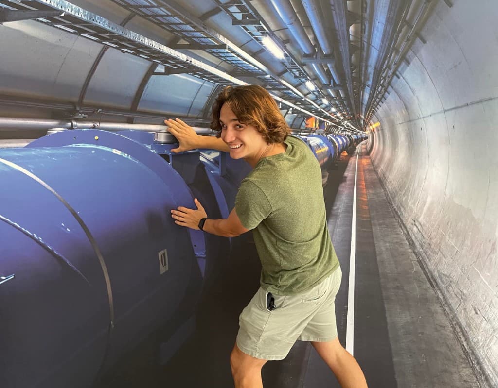 A founder smiling beside a large blue beam pipe in a long underground research tunnel with industrial lighting.